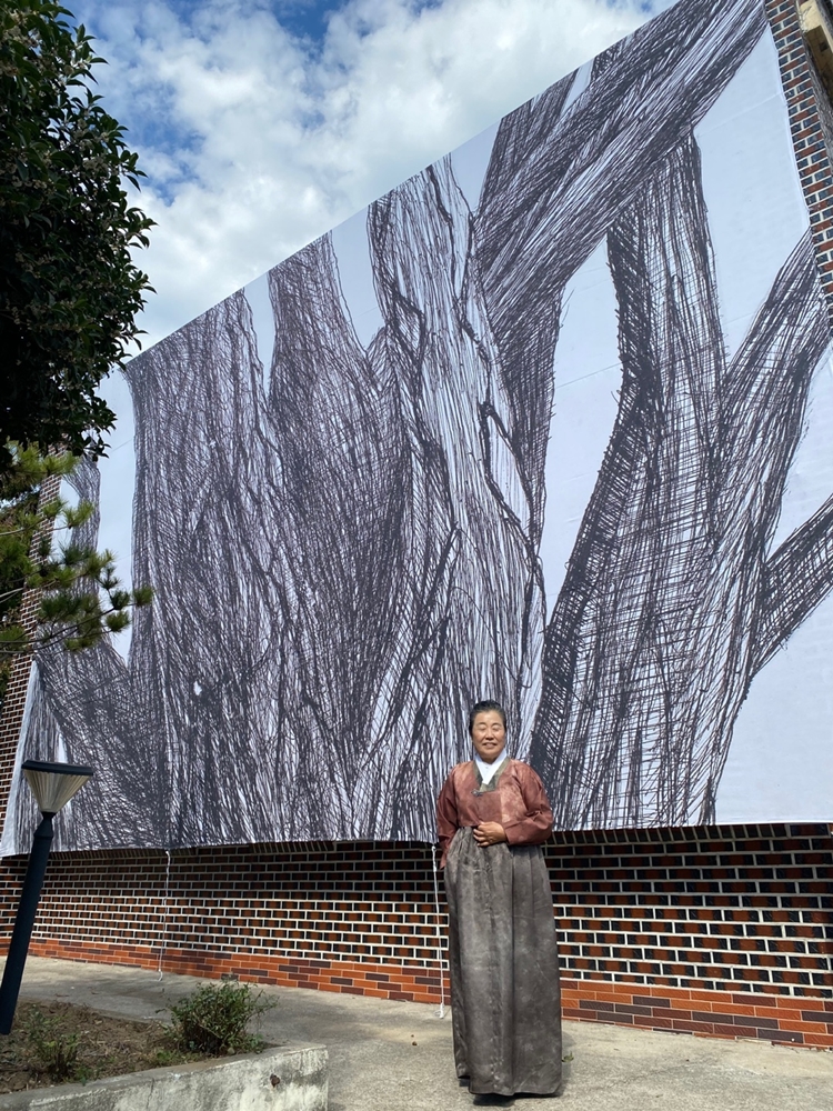 ▲ 이나경 작가의 최근작인 옥수수드로잉 작품이 에그갤러리 외벽에 걸려 있다. 작품 앞에 서 있는 이나경 작가.